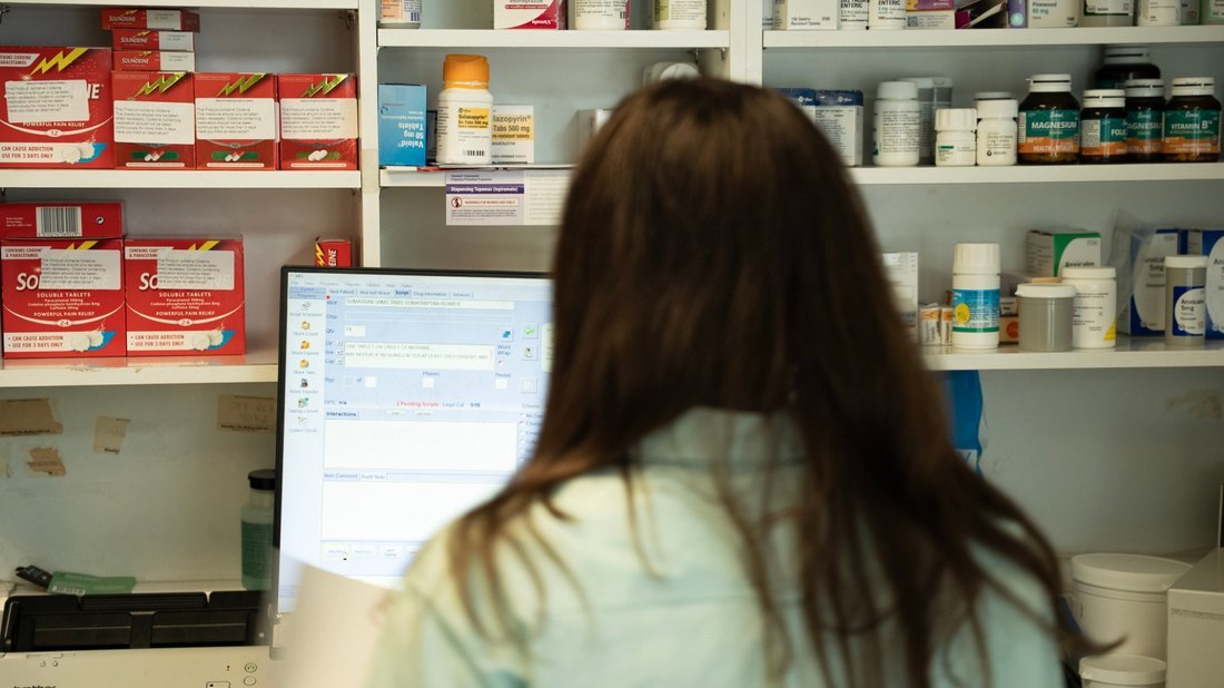 A pharmacist at the dispensary screen, shelves of stock filling the wall in front of them.